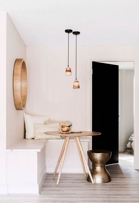 a minimalist breakfast nook with a white corner bench, a stained table, a metal brass stool, pendant lamps and a mirror in a wooden frame