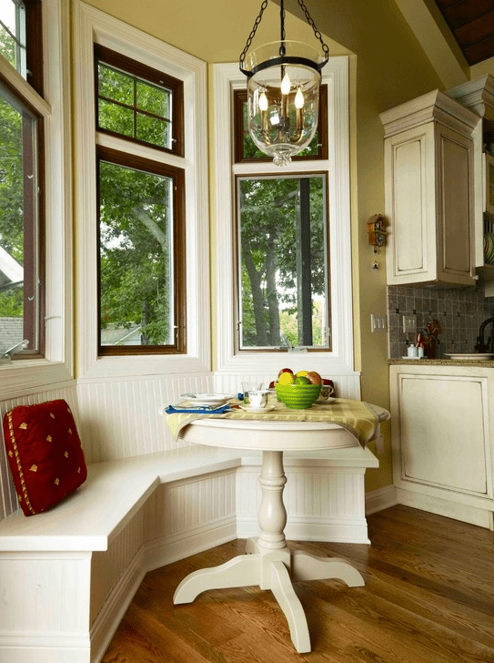 A neutral, country-style breakfast nook with built-in banquette seating, a small table, some pillows, and a pendant lamp