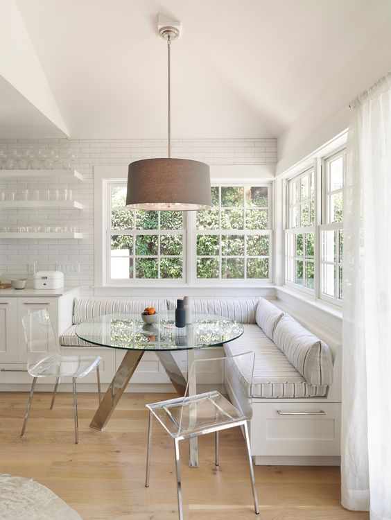 a neutral, country-style breakfast nook with large corner banquette seating, a glass table and acrylic chairs, and a hanging lamp