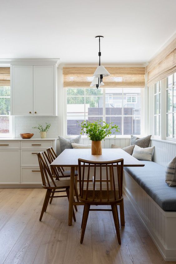 A calm, modern farmhouse breakfast nook with a sitting area, a stained table and chairs, a hanging lamp and some pillows