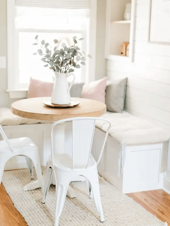 A white farmhouse breakfast nook with a built-in bench with storage, a round table, white metal chairs and built-in niches for storage