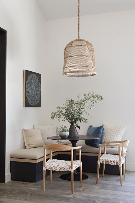 an earthy breakfast nook with a black corner bench and cushions, a black table and wicker chairs, a pendant lamp and some greenery