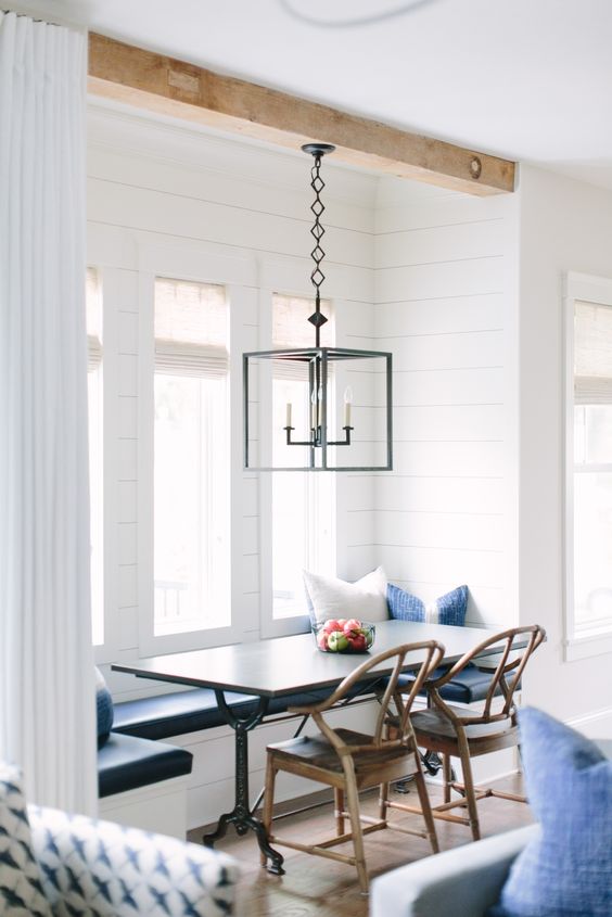 A coastal and rustic breakfast nook by the window with a dark table, rustic chairs, a hanging lamp and some blue and white pillows