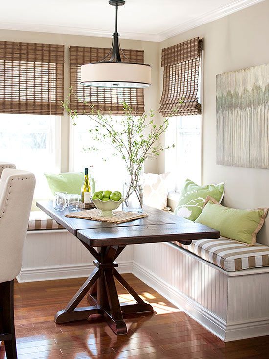 A green and brown farmhouse style breakfast nook with a sitting area, a dark stained table and various pillows, woven lampshades and green branches