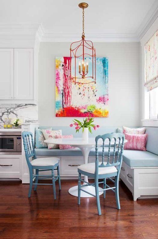 A pastel breakfast nook with an eye-catching piece of art, a blue corner cushion bench, blue chairs and some flowers is beautiful