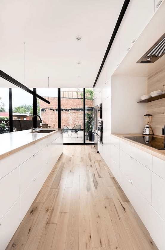 a cozy, minimalist galley kitchen in white with light wooden worktops and a floor and back wall, with glazed walls for more light