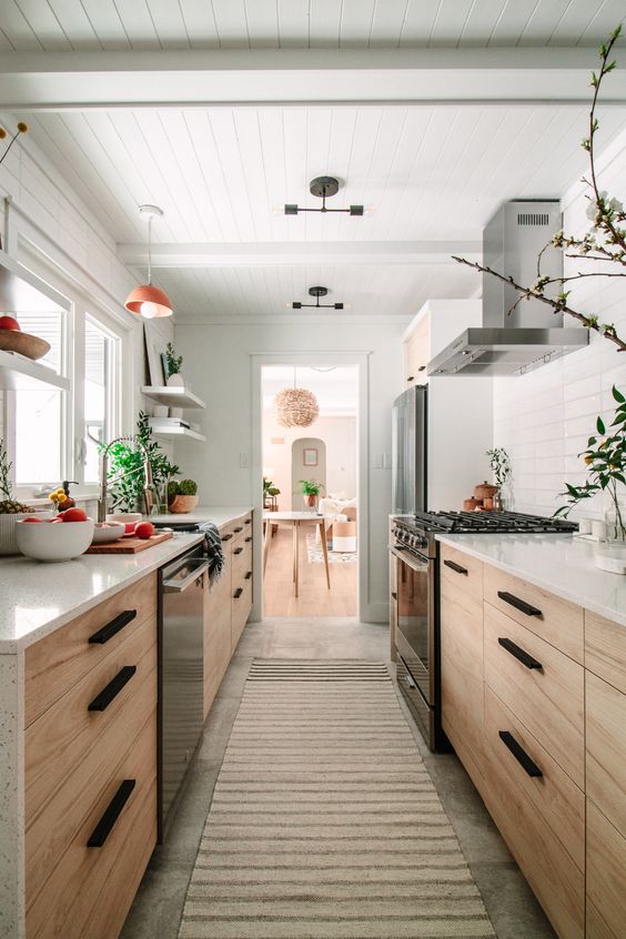 A modern, stained country style galley kitchen with white stone countertops and a white tile backsplash, lots of greenery and lots of light