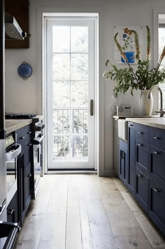 An inviting navy blue farmhouse kitchen with navy blue cabinets and wood countertops, a hardwood floor and lots of natural light through the door
