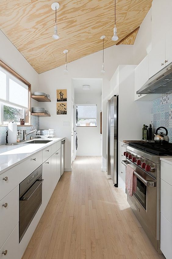 a modern white galley kitchen with a plywood ceiling with hanging lamps, a wooden floor and a blue tile backsplash