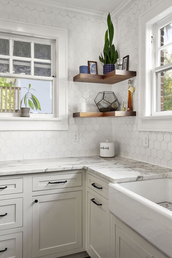 A beautiful neutral kitchen with shaker cabinets, white stone countertops, a white hexagon tile backsplash and corner shelves