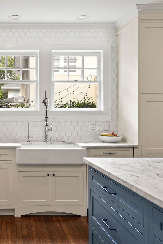 A cream vintage kitchen with white stone countertops, white hexagon tiles on the wall and a blue kitchen island