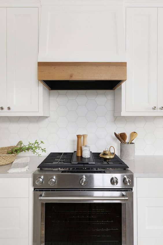 a sleek white kitchen with shaker cabinets, a wood-edged range hood, and a white hexagonal tile backsplash and neutral countertops