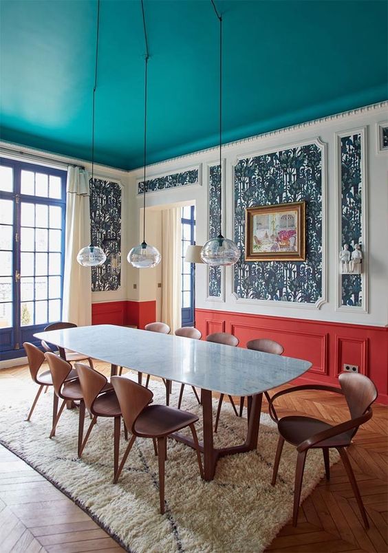a bright dining room with a turquoise ceiling, bold navy wallpaper and red paneling, a dining table and plywood chairs, and hanging lamps